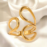 Offener goldfarbener Ring mit organischen, geschwungenen Linien in S-Form, fotografiert auf weißem, weich gefaltetem Satin. Das Licht bringt den warmen Glanz und die glatte Oberfläche des Designs deutlich hervor.