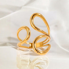 Offener goldfarbener Ring mit organischen, geschwungenen Linien in S-Form, fotografiert auf weißem, weich gefaltetem Satin. Das Licht bringt den warmen Glanz und die glatte Oberfläche des Designs deutlich hervor.