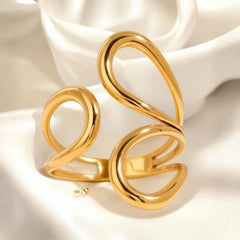 Offener goldfarbener Ring mit organischen, geschwungenen Linien in S-Form, fotografiert auf weißem, weich gefaltetem Satin. Das Licht bringt den warmen Glanz und die glatte Oberfläche des Designs deutlich hervor.