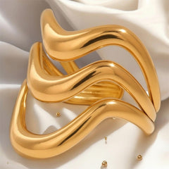 Dreifach übereinanderliegende, wellenförmige goldene Ringe auf einem sanft gefalteten weißen Satinhintergrund. Das Licht betont den warmen Goldglanz und die geschmeidige, organische Form der Ringe.“