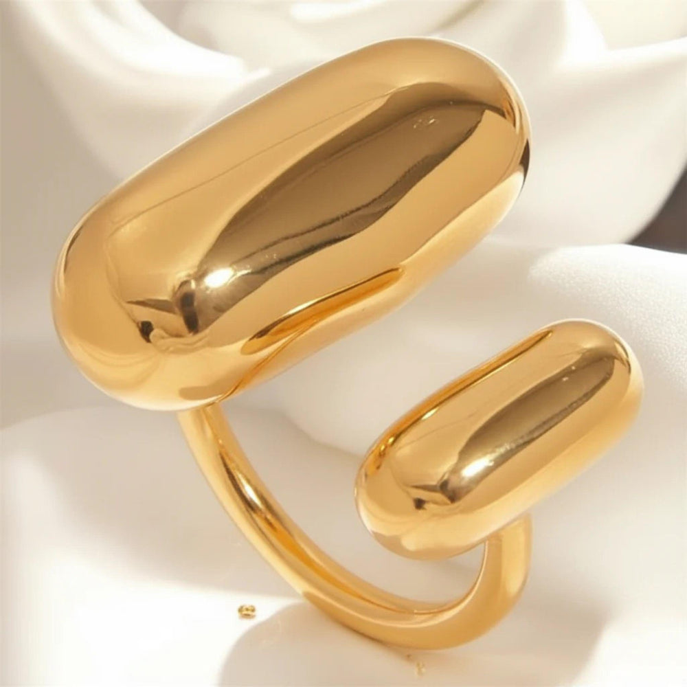Offener goldfarbener Ring mit zwei polierten, ovalen Kapsel-Elementen in unterschiedlicher Größe, arrangiert auf weich gefaltetem weißen Satin. Das warme Licht verstärkt den spiegelnden Glanz und die glatte, moderne Form des Rings.