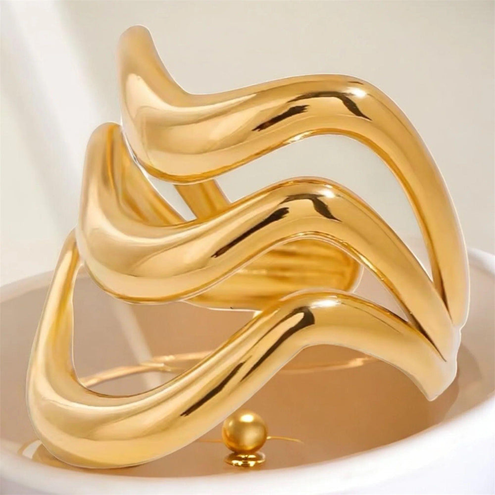 Dreifach übereinanderliegende, wellenförmige goldene Ringe auf einem sanft gefalteten weißen Satinhintergrund. Das Licht betont den warmen Goldglanz und die geschmeidige, organische Form der Ringe.“