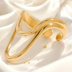 Offener, goldfarbener Statement-Ring mit einer asymmetrischen, geschwungenen V-Form, fotografiert auf weich fallendem weißen Satin. Das warme Licht betont den Glanz und die fließende Linienführung des Designs.